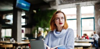 Cheerful young beautiful redhaired woman in glasses using her phone touchpad and notebook while sitting at her working place on cafe with cup of coffee
