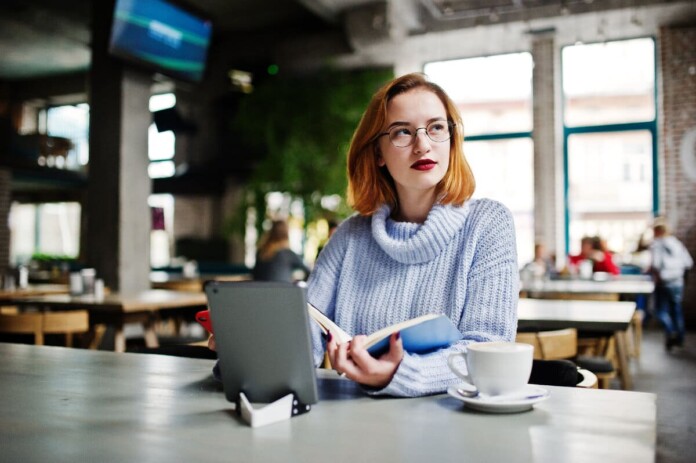 Cheerful young beautiful redhaired woman in glasses using her phone touchpad and notebook while sitting at her working place on cafe with cup of coffee